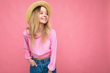 Photo of pretty young female person wearing pink blouse and beige summer hat isolated over pink background looking to the side smilimg and having funの写真素材