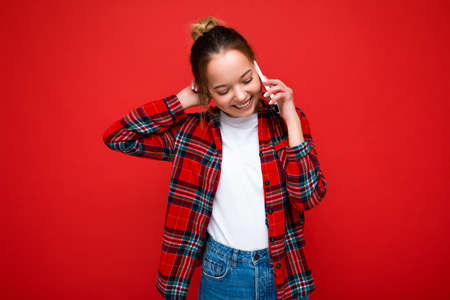 Attractive cute positive young blonde woman standing isolated over red wall wearing casual red shirt and white t-shirt talking on mobile phone looking downの写真素材