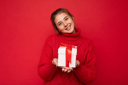 Beautiful happy young brunette woman isolated over colourful background wall wearing stylish casual clothes holding gift box and looking at cameraの写真素材