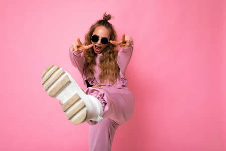 Photo of happy positive young attractive blonde curly woman with sincere emotions wearing trendy pink sportswear and sunglasses isolated on pink background with copy space and having funの写真素材