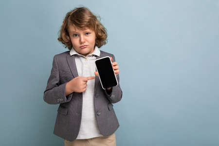 Handsome boy with curly hair wearing suit holding phone isolated over blue background looking at...