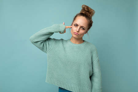 Portrait of young sad sorrowful emotional attractive blonde woman with sincere emotions wearing casual blue jersey isolated over blue background with empty space and making finger pistol near headの写真素材