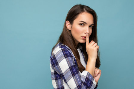 Young serious beautiful brunet woman with sincere emotions wearing trendy check shirt standing isolated on blue background with empty space and showing shhh gesture. Keep secret conceptの写真素材