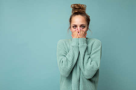 Portrait of young shocked nice winsome blond woman with sincere emotions wearing trendy blue sweater isolated over blue background with free space and covering mouthの写真素材