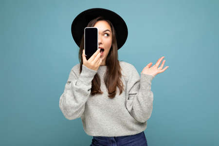 Amazed Beautiful positive woman wearing black hat and grey sweater holding mobilephone showing smartphone isolated on background looking to the side with open mouthの写真素材