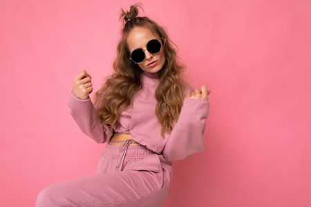 Shot of charming happy young dark blonde curly woman isolated over pink background wall wearing casual pink sport clothes and stylish sunglasses looking at camera and dancingの写真素材