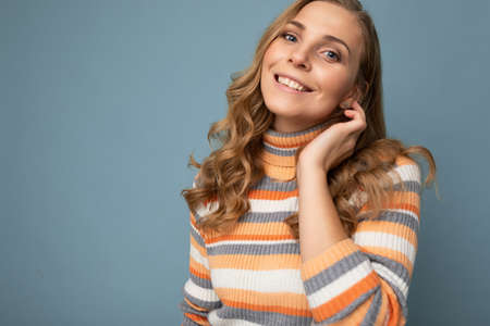 Portrait of young winsome attractive happy smiling blonde woman with wavy-hair wearing striped sweater isolated over blue background with empty spaceの写真素材