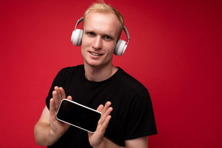 Handsome young blonde man wearinng casual black t-shirt and headphones listening to podcast holding...