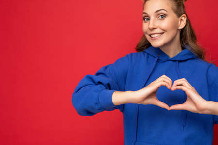 Portrait of young happy positive beautiful blonde woman with sincere emotions wearing trendy bright blue hoodie isolated on red background with copy space and showing heart shape with handsの写真素材