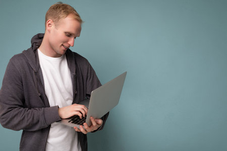 Side profile photo shot of handsome blonde man holding computer laptop typing on keyboard wearing white t-shirt and grey sweater looking at netbook isolated over blue backgroundの写真素材