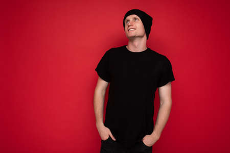 Handsome positive self-confident young man standing isolated over red background wall wearing casual black t-shirt for mockup and black hat and looking to the sideの写真素材
