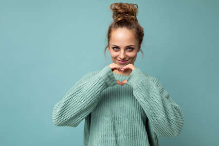 Portrait of young happy positive smiling beautiful blonde woman with sincere emotions wearing casual blue pullover isolated on blue background with copy space and gesturing with hands heart shapeの写真素材