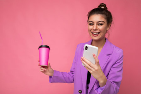 Young positive laughing woman isolated over pink background with copy space holding coffee to take away and a mobile phoneの写真素材