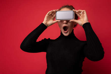 Photo of beautiful shocked young brunette woman wearing black sweater standing isolated over red background showing mobile phone with empty screen for mockup with open mouthの写真素材