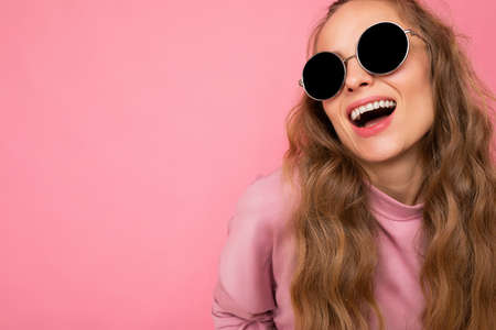 Closeup shot of beautiful happy young dark blonde curly woman isolated over pink background wall wearing casual pink sport clothes and stylish sunglasses looking at cameraの写真素材