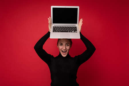Close-up portrait of Beautiful smiling happy young woman with gathered brunet hair holding computer laptop with empty monitor screen with mock up wearing black longsleeve looking at cameraの写真素材