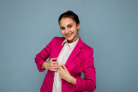 Photo portrait of young attractive beautiful positive smiling brunette woman with sincere emotions wearing casual white shirt and stylish pink jacket isolated over blue background with copy spaceの写真素材