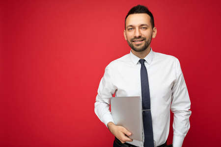 Handsome smiling brunet man holding laptop computer looking at camera in white shirt and tie on isolated red backgroundの写真素材