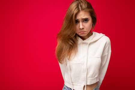 Sorrowful young attractive dark blonde woman with sincere emotions isolated on background wall with copy space wearing casual white hoodie. Sad conceptの写真素材