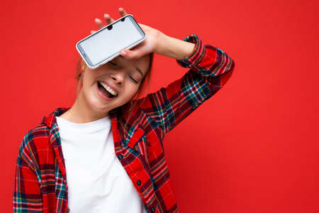 Photo of jouful beautiful positive young blonde woman standing isolated over red wall wearing casual red shirt and white t-shirt showing mobile phone with empty screen for mockupの写真素材