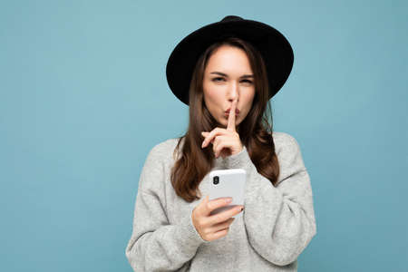 young woman wearing black hat and grey sweater holding smartphone looking at camera showing shhh gesture isolated on backgroundの写真素材