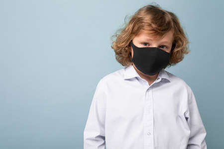 Photo of handsome curly blonde little boy in medical black mask standing isolated over blue background.の写真素材