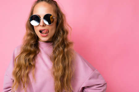 Pretty positive sexy young blonde curly woman isolated over pink background wall wearing casual pink sport clothes and stylish sunglasses looking at cameraの写真素材