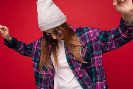 Fantastic photo shot of beautiful young dark blonde woman isolated over red background wearing hipster purple casual shirt grey hat and stylish sunglasses looking down and dancingの写真素材