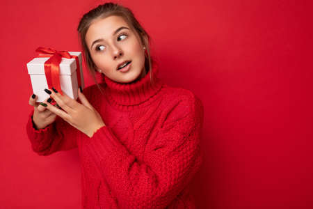 Photo of pretty positive thoughtful brunette young woman isolated over red background wall wearing red sweater holding white gift box with red ribbon and looking to the sideの写真素材