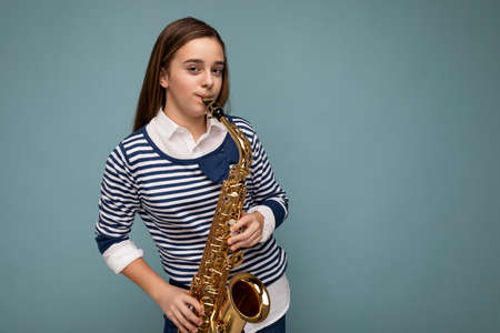 Shot of pretty positive happy brunet little lady wearing stylish striped longsleeve standing isolated over blue background wall playing saxophone looking at cameraの写真素材