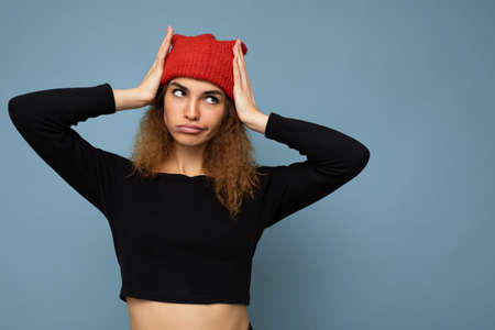 Photo of young pretty amusing positive brunette woman curly with sincere emotions wearing stylish black crop top and red hat isolated on blue background with copy space and grimacingの写真素材