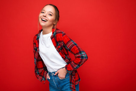 Young beautiful woman in stylish red hipster shirt and casual white t-shirt for mockup. Positive female shows facial emotions. Funny model isolated on red background with free spaceの写真素材