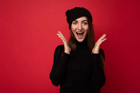 Photo of young positive surprised happy attractive brunette woman with sincere emotions wearing black roll-neck sweater and black hat isolated over red background with copy space. Shock conceptの写真素材