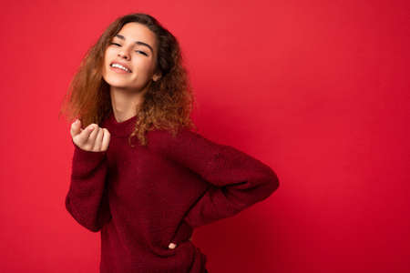 Portrait of young positive happy attractive brunette curly woman with sincere emotions wearing casual red sweater isolated on red background with copy spaceの写真素材