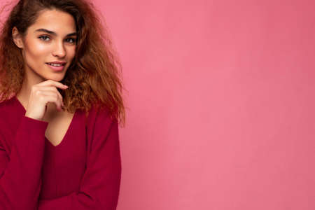 Portrait of young positive thoughtful beautiful brunette curly woman with sincere emotions wearing trendy pink pullover isolated on pink background with copy spaceの写真素材