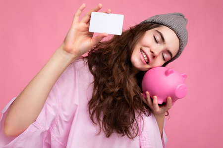 Charming smiling happy joyful young curly brunette woman wearing pink shirt and gray hat isolated on pink background with free space and holding pink pig money box and credit card for mockupの写真素材