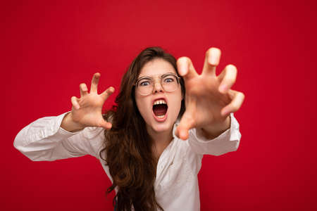 Portrait of young emotional attractive brunette woman with sincere emotions wearing casual white shirt and optical glasses isolated on red background with free space and doing claw gesture as cat with agressive and sexy expression. Roar concept.の写真素材