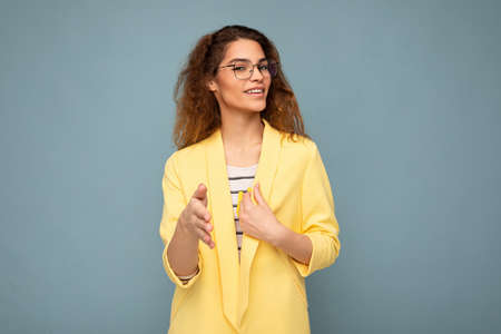 Photo of young positive happy attractive brunette curly woman with sincere emotions wearing stylish yellow jacket and optical glasses isolated on blue background with copy spaceの写真素材