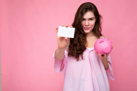 Portrait of beautiful positive cheerful cute smiling young brunette woman in stylish shirt isolated on pink background with copy space and holding pink pig moneybox and credit card for mockupの写真素材