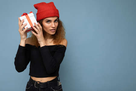 Photo shot of beautiful concentrated thinking adult dark blonde curly woman isolated over blue background wall wearing black crop top and red hat holding gift box looking at cameraの写真素材