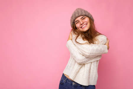 Shot of beautiful happy positive smiling young brunette woman isolated over pink background wall wearing knitted white sweater and grey warm hat and having funの写真素材
