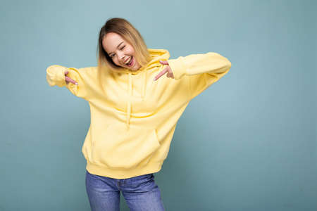 Photo shot of young beautiful cute happy blonde woman wearing stylish yellow hoodie isolated over blue background with copy spaceの写真素材