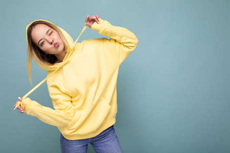 Photo shot of young beautiful cute happy blonde woman wearing stylish yellow hoodie isolated over blue background with copy spaceの写真素材