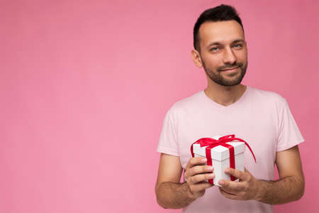 Handsome confident brunet man holding white gift box with red ribbon with surprisepresent in pink...