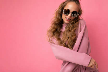 Photo shot of young beautiful attractive cute cool positive curly blonde woman wearing trendy and stylish pink sport suit and sunglasses isolated over pink background with copy space for textの写真素材