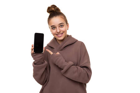 Photo of a beautiful cute girl with dark hair and a bun in a brown hoodie with a hood holding a mobile phone with a blank black screen isolated on a white background with free space for textの写真素材
