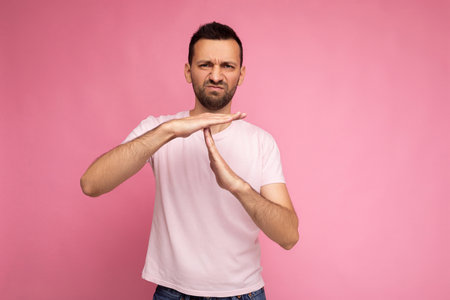 Photo of handsome unhappy angry dissatisfied brunet man in casual pink t-shirt isolated on pink background with copy space for text and showing time out and stop gesture with handsの写真素材