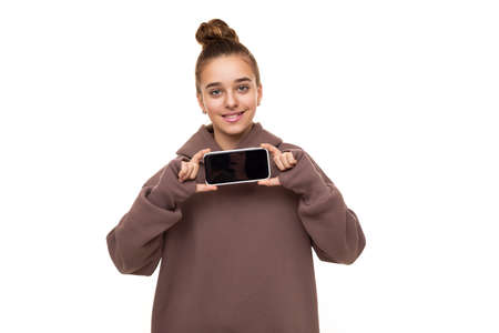 Photo of a beautiful cute girl with dark hair and a bun in a brown hoodie with a hood holding a mobile phone with a blank black screen isolated on a white background with free space for textの写真素材