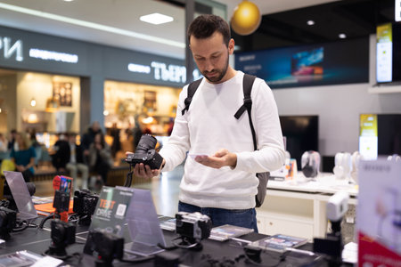 Minsk, Belarus - Nov 27, 2021: Photo of an adult handsome thinking brunet man with stubble in a white sweatshirt chooses a camera in an electronic store in a shopping mall. Shop and price conceptのeditorial素材