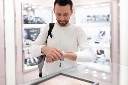 Photo of an adult handsome brunette man in a white sweatshirt chooses and tries on a stylish wrist...